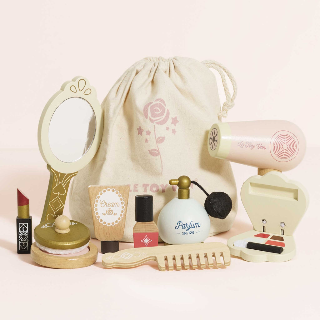 A collection of pastel-colored toy cosmetics, including perfume, lipstick, a mirror, and a hairbrush, arranged with a drawstring bag. The scene is playful and whimsical.
