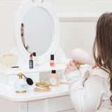 Child using a toy wooden hair dryer at a white vanity table set with play cosmetics.