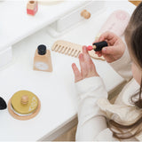Child playing with a wooden toy lipstick at a white vanity table with beauty accessories.