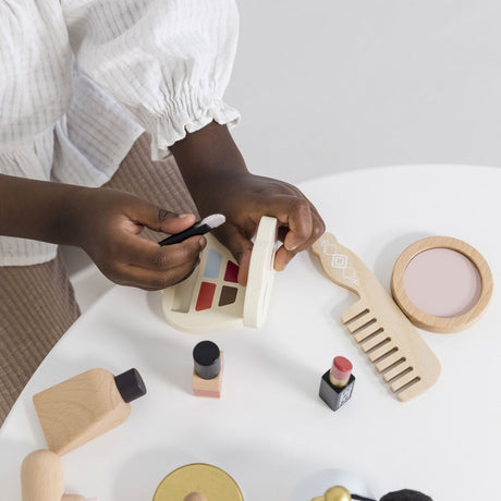 Child using a brush with a wooden toy makeup palette at a white table.