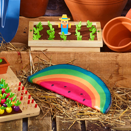 Vilac rainbow watermelon pouch resting on straw next to wooden garden toys and flower pots.