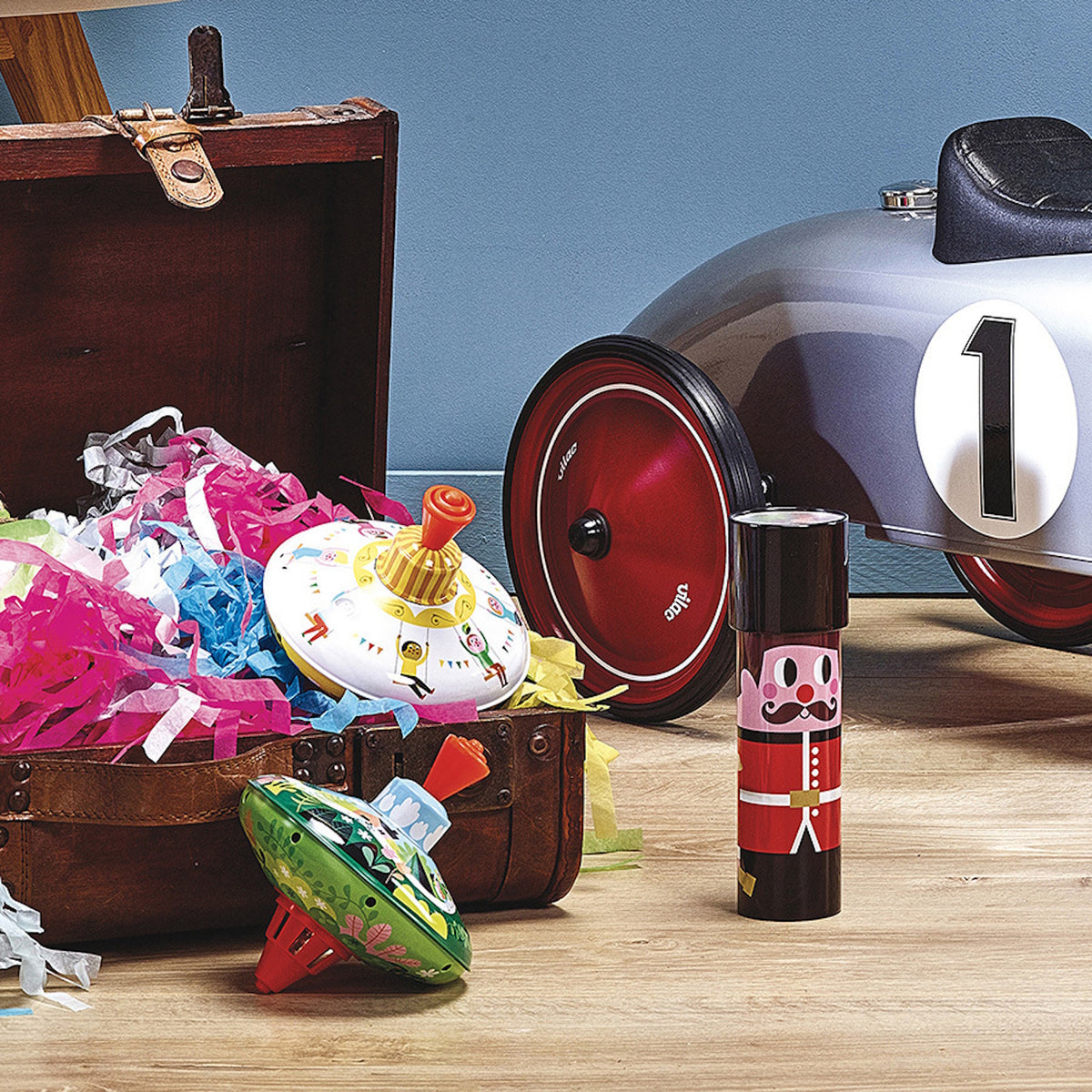 Red and black metal guardsman kaleidoscope toy standing beside a toy car and treasure chest filled with colorful paper in a lifestyle room view.