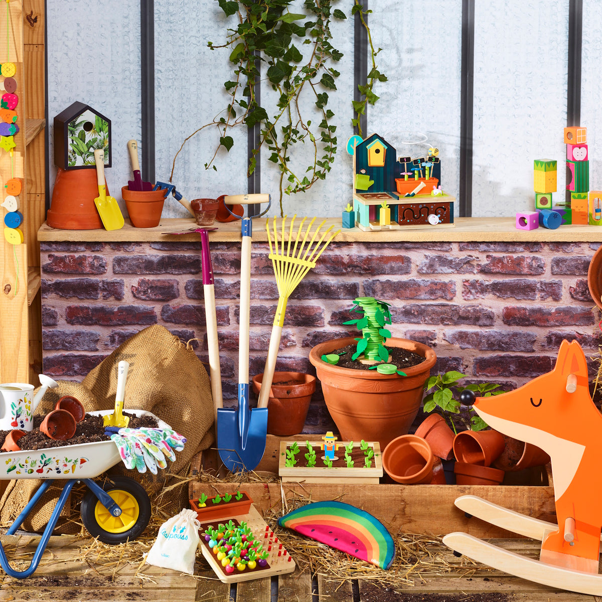 Colorful wooden garden toys including a wheelbarrow, rake, shovel, and fox rocker displayed against a brick wall.