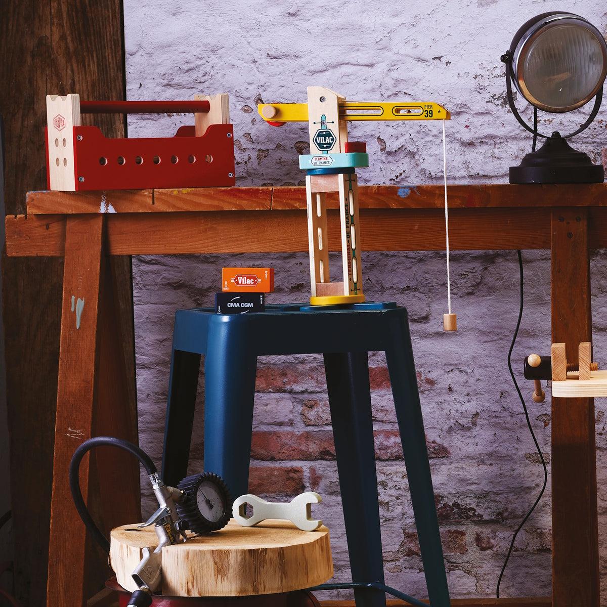 Vilac wooden toy crane and tool box on a workbench with a blue stool and tools in a rustic workshop setting.