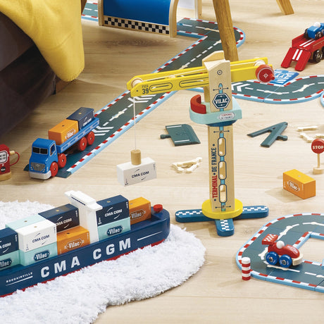 Wooden toy harbor set with a crane, CMA CGM container ship, truck, and road tracks on a light wood floor.