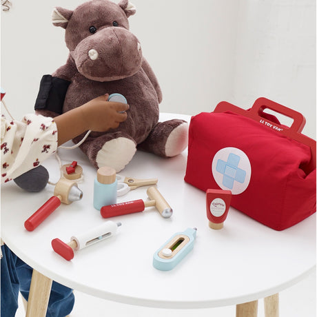 Child using a wooden play stethoscope on a stuffed hippo next to a red medical kit bag.