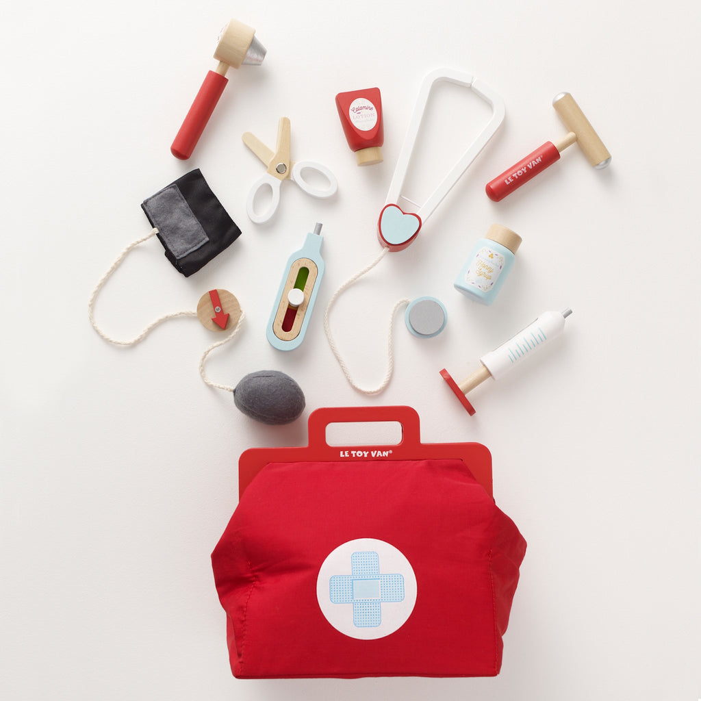 A vibrant red toy medical bag with a white cross symbol, surrounded by wooden toy instruments like a syringe, scissors, a stethoscope, and medicine bottles.