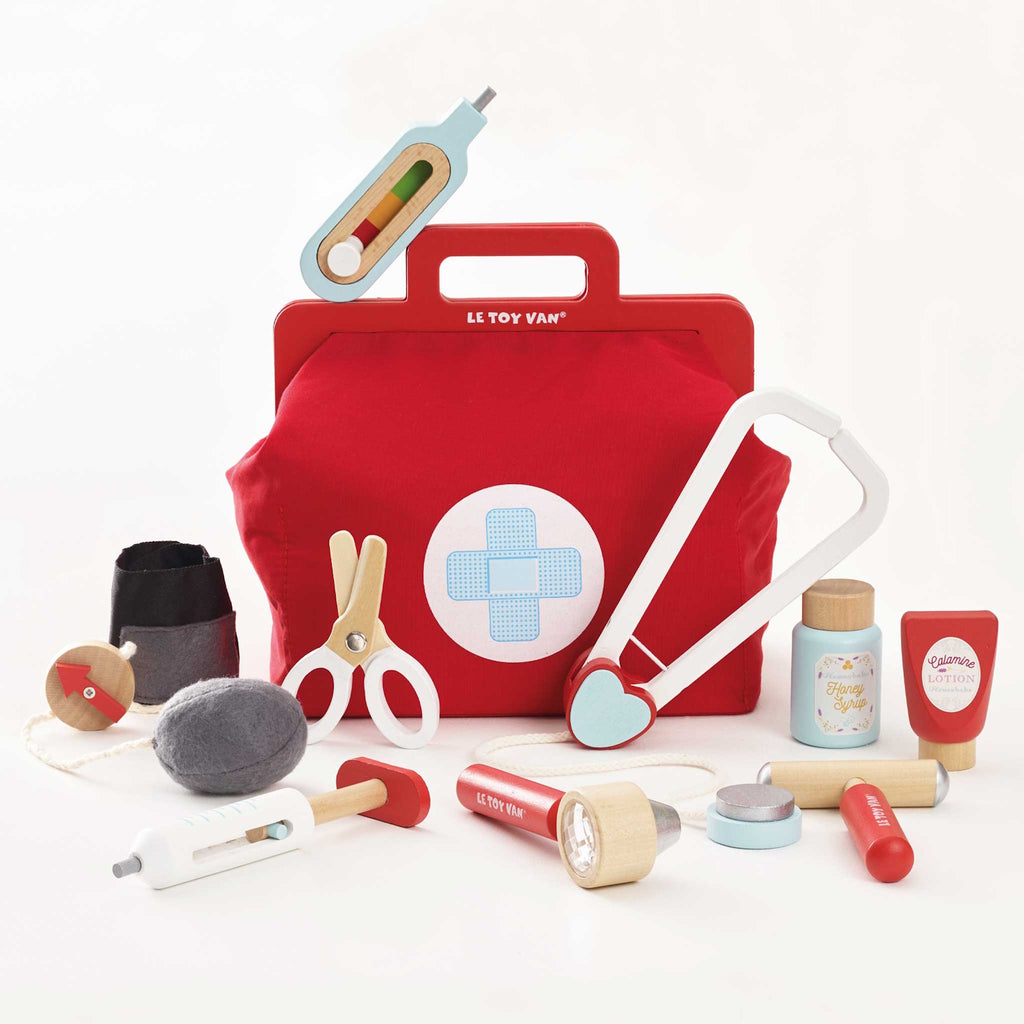 A vibrant red toy medical bag with a white cross symbol, surrounded by wooden toy instruments like a syringe, scissors, a stethoscope, and medicine bottles.
