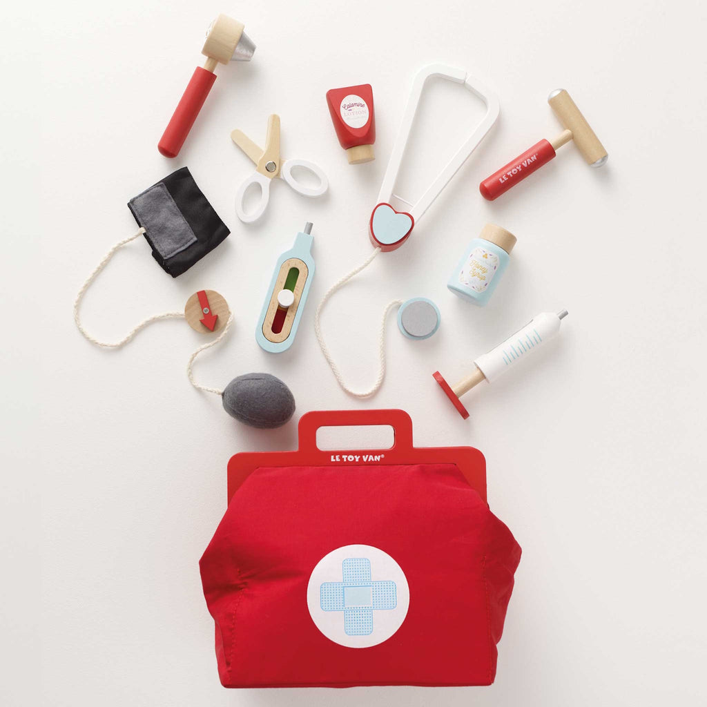 A vibrant red toy medical bag with a white cross symbol, surrounded by wooden toy instruments like a syringe, scissors, a stethoscope, and medicine bottles.