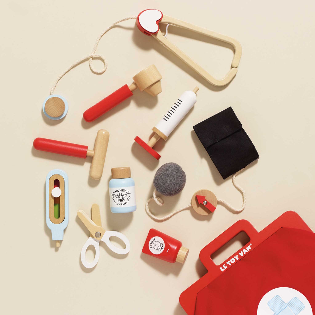 A vibrant red toy medical bag with a white cross symbol, surrounded by wooden toy instruments like a syringe, scissors, a stethoscope, and medicine bottles.