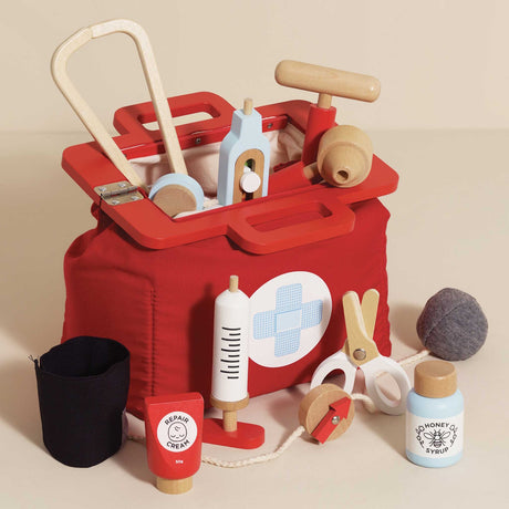 Open red wooden doctor bag filled with medical play tools, with additional accessories set in front against a cream background.