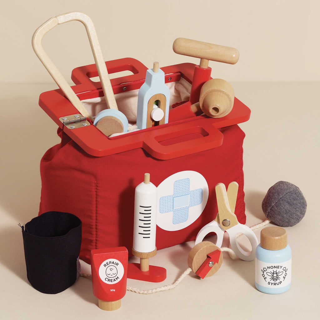 A vibrant red toy medical bag with a white cross symbol, surrounded by wooden toy instruments like a syringe, scissors, a stethoscope, and medicine bottles.