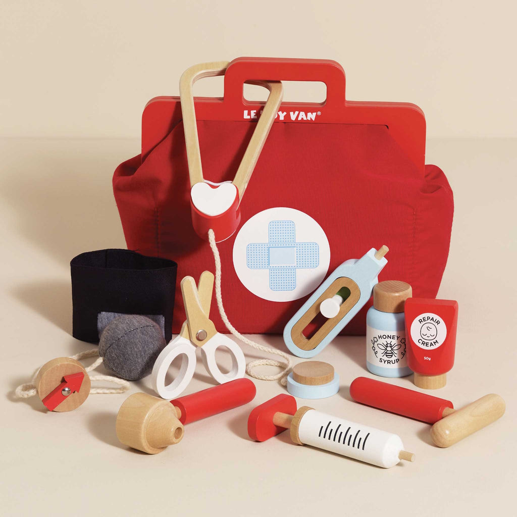 A vibrant red toy medical bag with a white cross symbol, surrounded by wooden toy instruments like a syringe, scissors, a stethoscope, and medicine bottles.