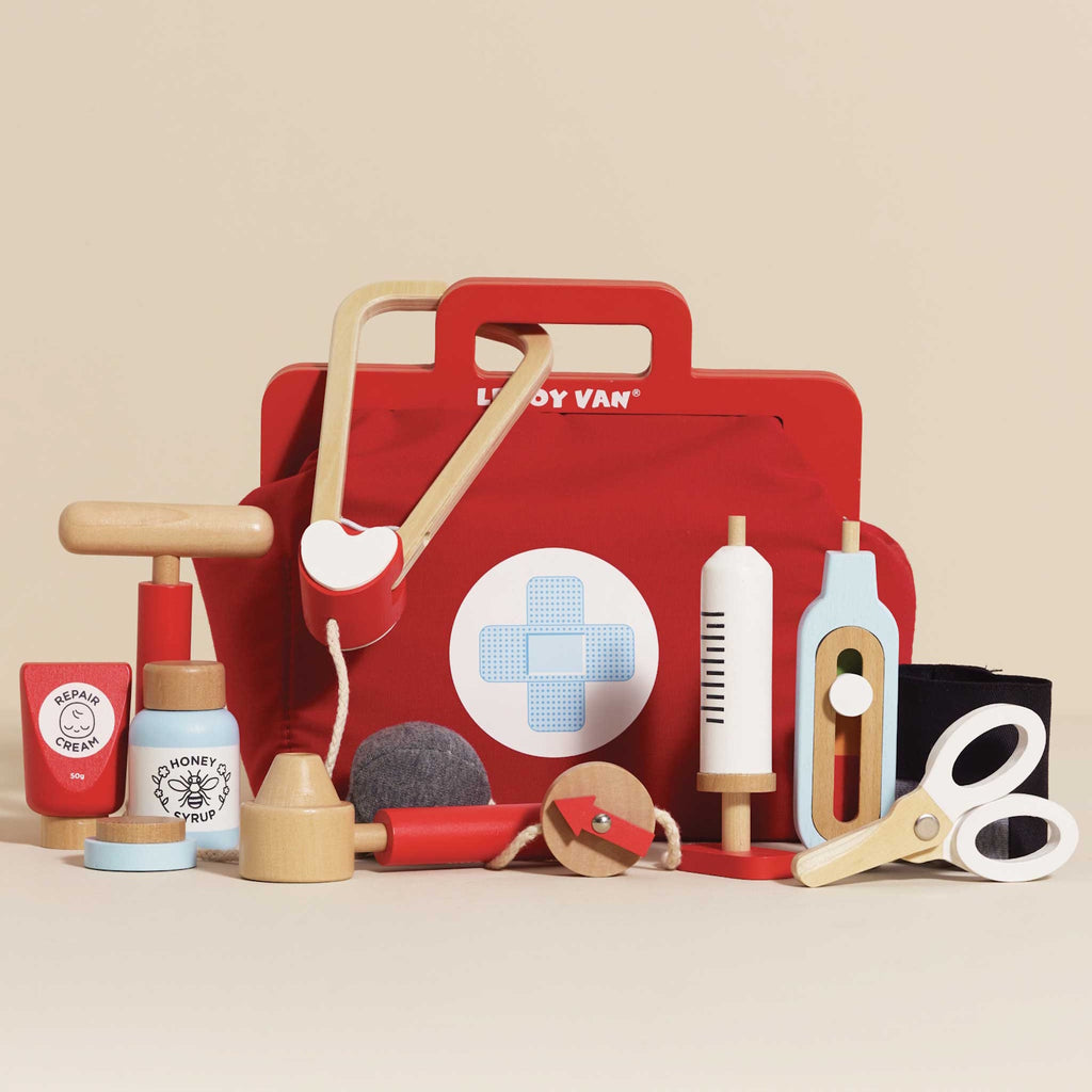 A vibrant red toy medical bag with a white cross symbol, surrounded by wooden toy instruments like a syringe, scissors, a stethoscope, and medicine bottles.