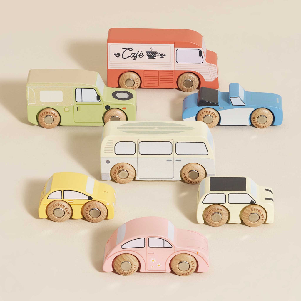 Colorful wooden toy vehicles on display include a red café truck, green SUV, blue sports car, yellow taxi, pink car, cream bus, and beige camper.