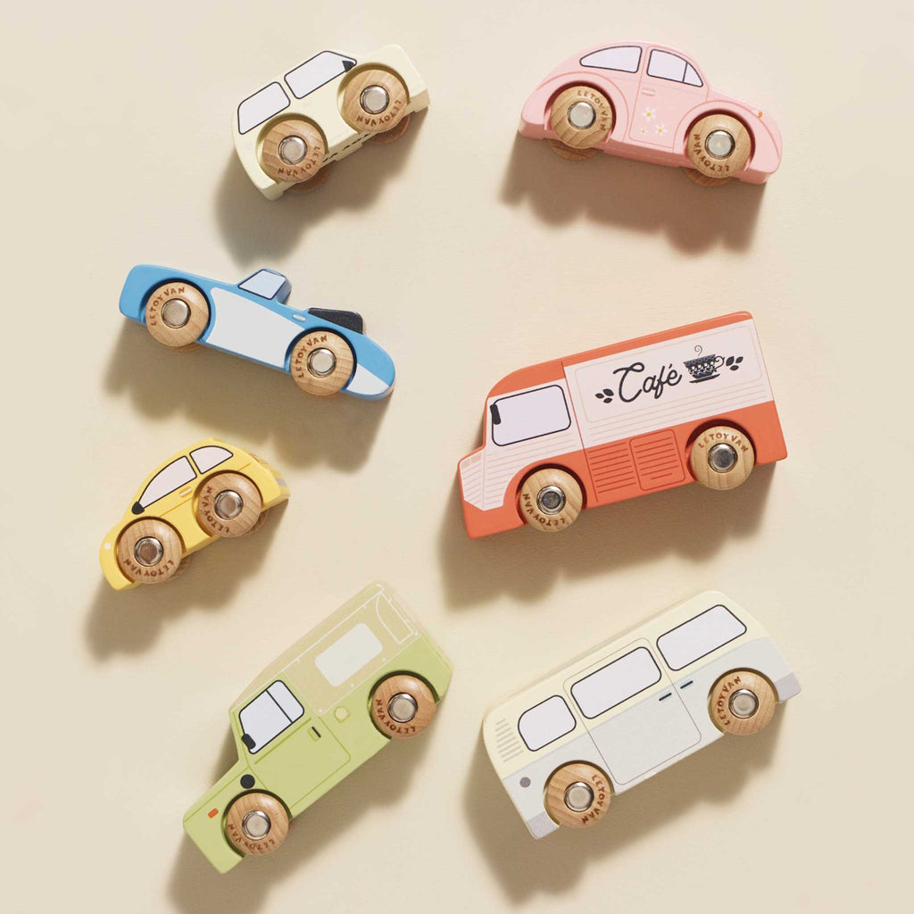 Colorful wooden toy vehicles on display include a red café truck, green SUV, blue sports car, yellow taxi, pink car, cream bus, and beige camper.