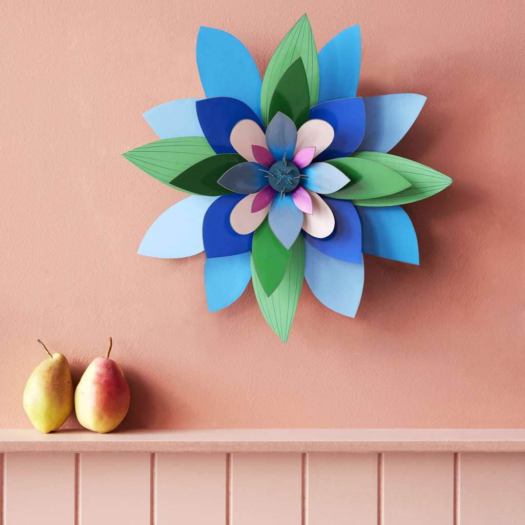A vibrant paper flower with blue, green, and pink petals adorns a peach wall. Below, two pears sit on a pink shelf, adding warmth and contrast.