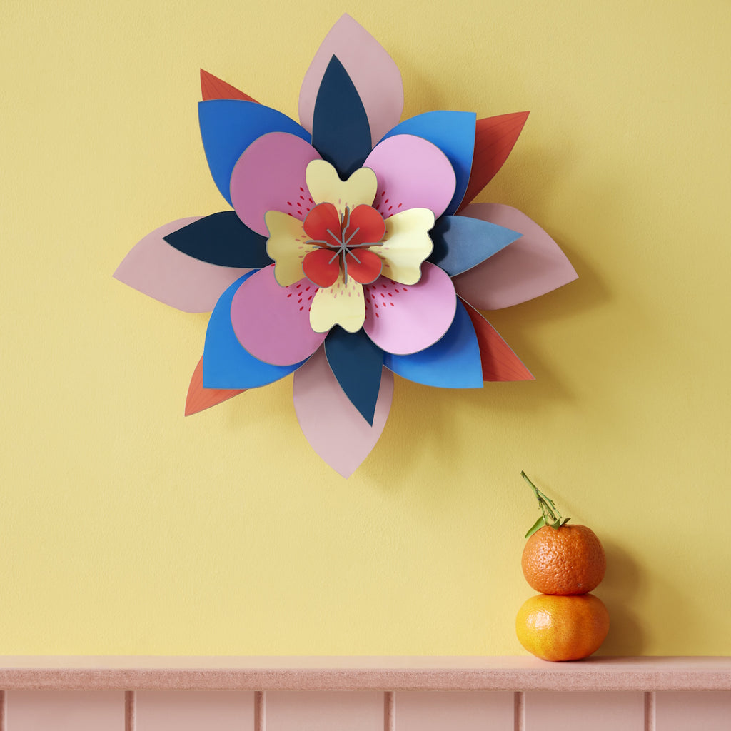Colorful paper flower with layered petals in blue, pink, and red on a yellow wall. Two stacked oranges sit on a light pink ledge below.