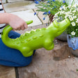 Green crocodile watering can used by a child to water white flowers in a garden.