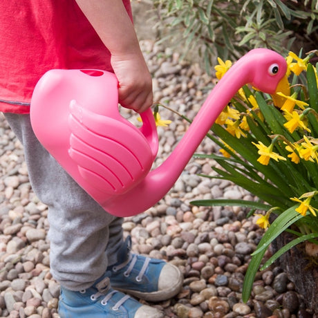 A child uses a pink flamingo-shaped watering can to water bright yellow daffodils in a pebble garden.