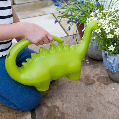 Green stegosaurus dinosaur watering can being used by a child to water white flowers in a garden.