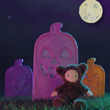 Plush doll in a brown werewolf suit sitting outdoors in front of pumpkin tombstones under a full moon.