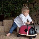 Toddler pushing a ladybird Wheely Bug ride on toy beside a lit Christmas tree and wrapped gifts.
