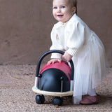 Smiling toddler in a white dress playing with a Wheely Bug ladybird ride-on toy.