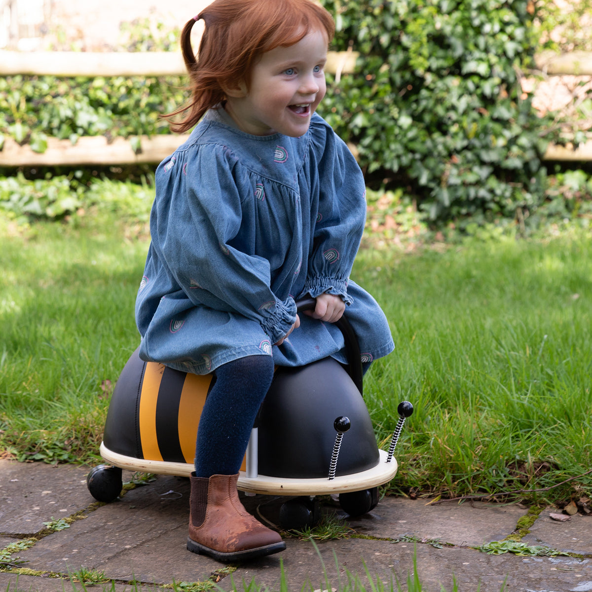 Happy toddler riding a Wheely Bug bee ride-on toy in a backyard garden.