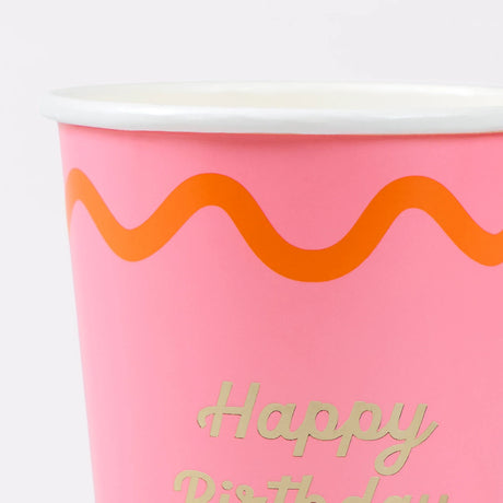 Close-up of a pink paper cup with an orange wavy line and gold text that reads "Happy Birthday".