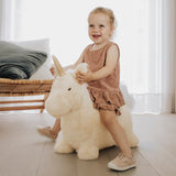Young girl playing on a plush white unicorn bouncy toy in a bright living room.