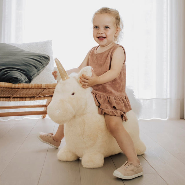 Young girl playing on a plush white unicorn bouncy toy in a bright living room.