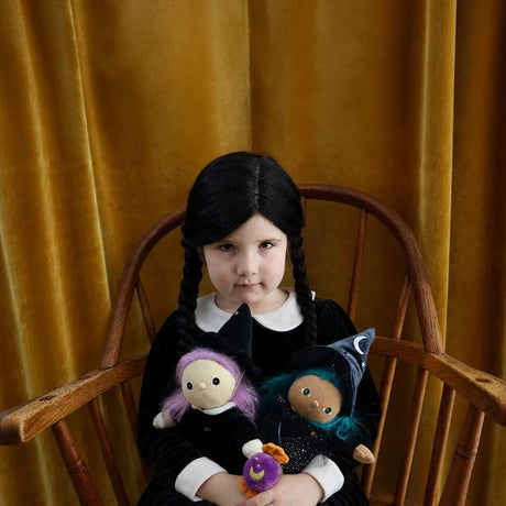 Child with dark braids sitting in a wooden chair holding two plush witch dolls.