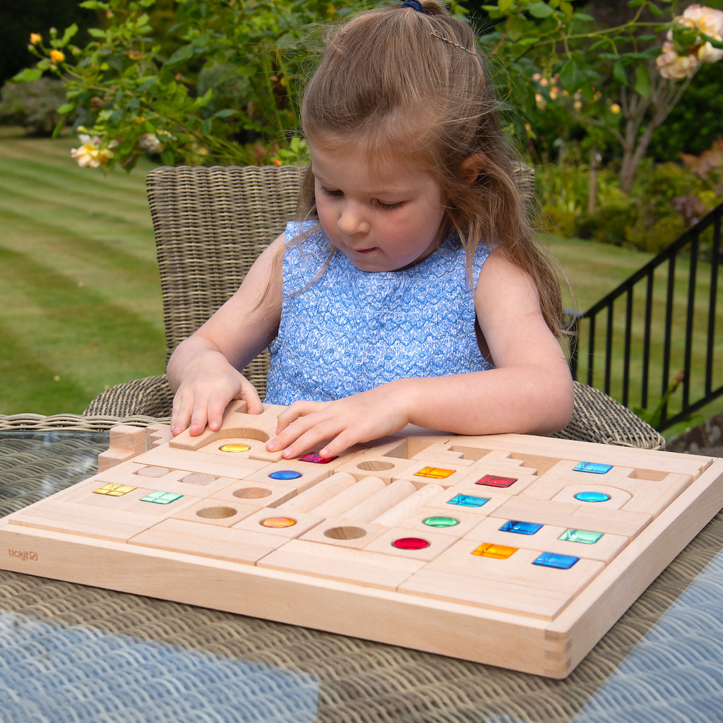 A young child builds a tower with wooden blocks featuring colorful transparent windows. The setting is bright and cheerful, promoting creativity and concentration.