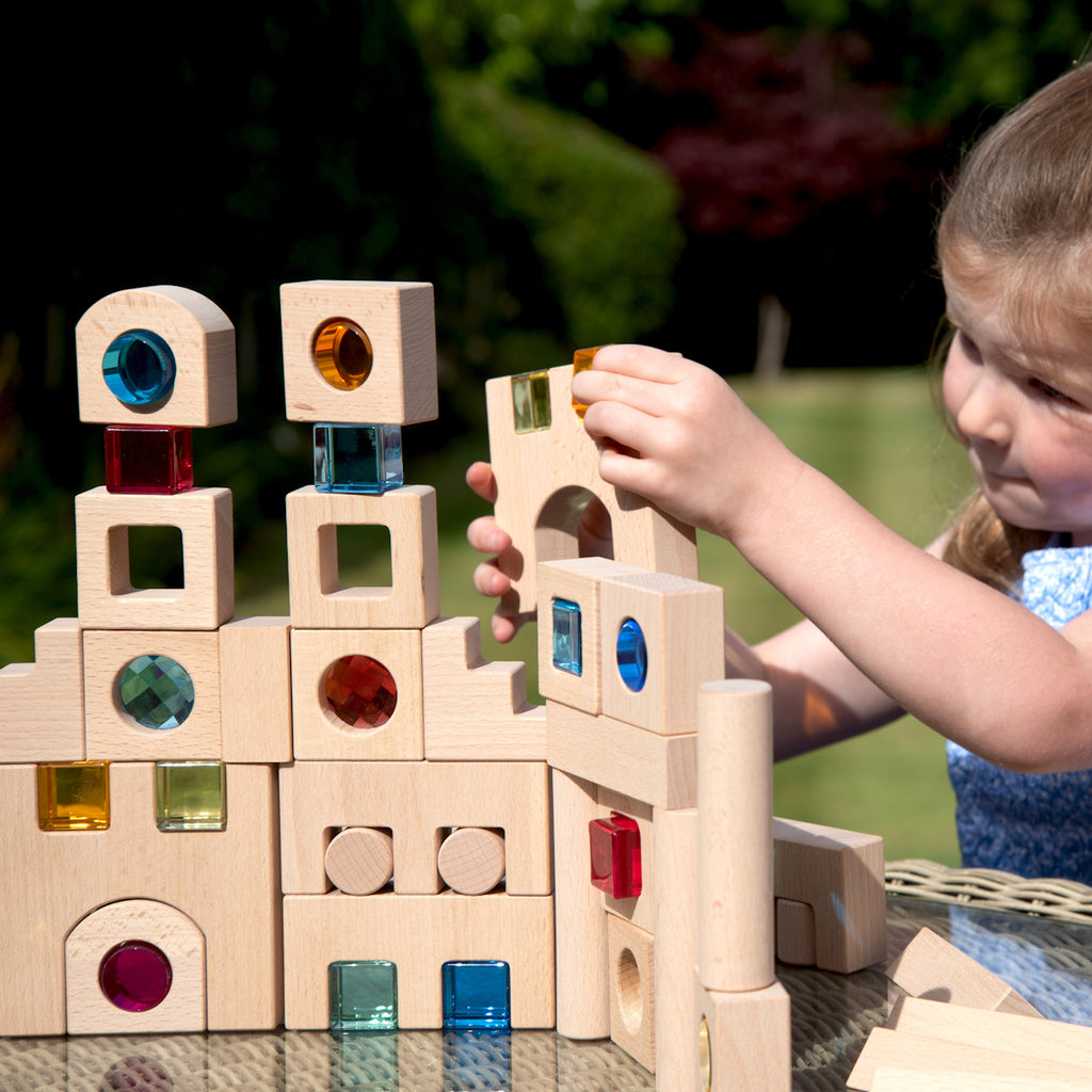 A young child builds a tower with wooden blocks featuring colorful transparent windows. The setting is bright and cheerful, promoting creativity and concentration.