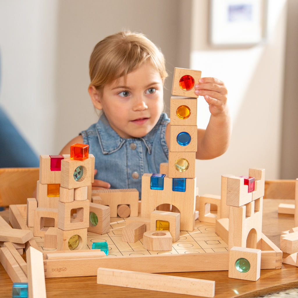 A young child builds a tower with wooden blocks featuring colorful transparent windows. The setting is bright and cheerful, promoting creativity and concentration.