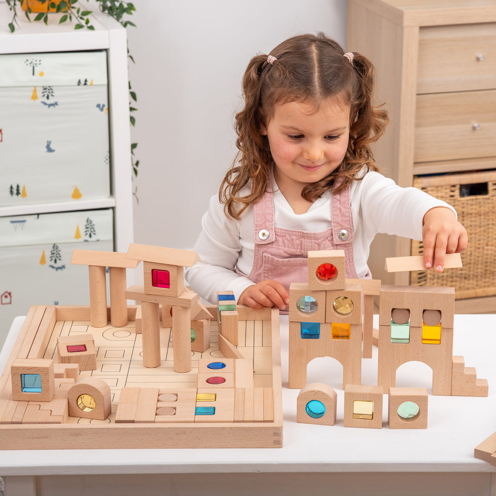 A young child builds a tower with wooden blocks featuring colorful transparent windows. The setting is bright and cheerful, promoting creativity and concentration.