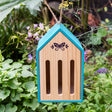 Teal wooden butterfly hotel hanging by a twine rope in a lush garden surrounded by green leaves and purple flowers.