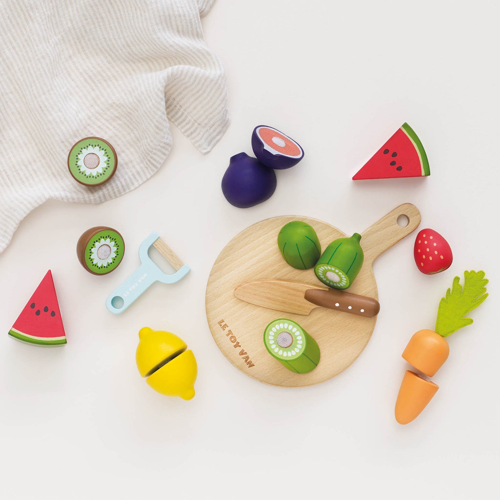 Wooden toy fruits and vegetables, including watermelon, kiwi, and carrot on a cutting board, with a toy knife and peeler, evoke playful cooking.