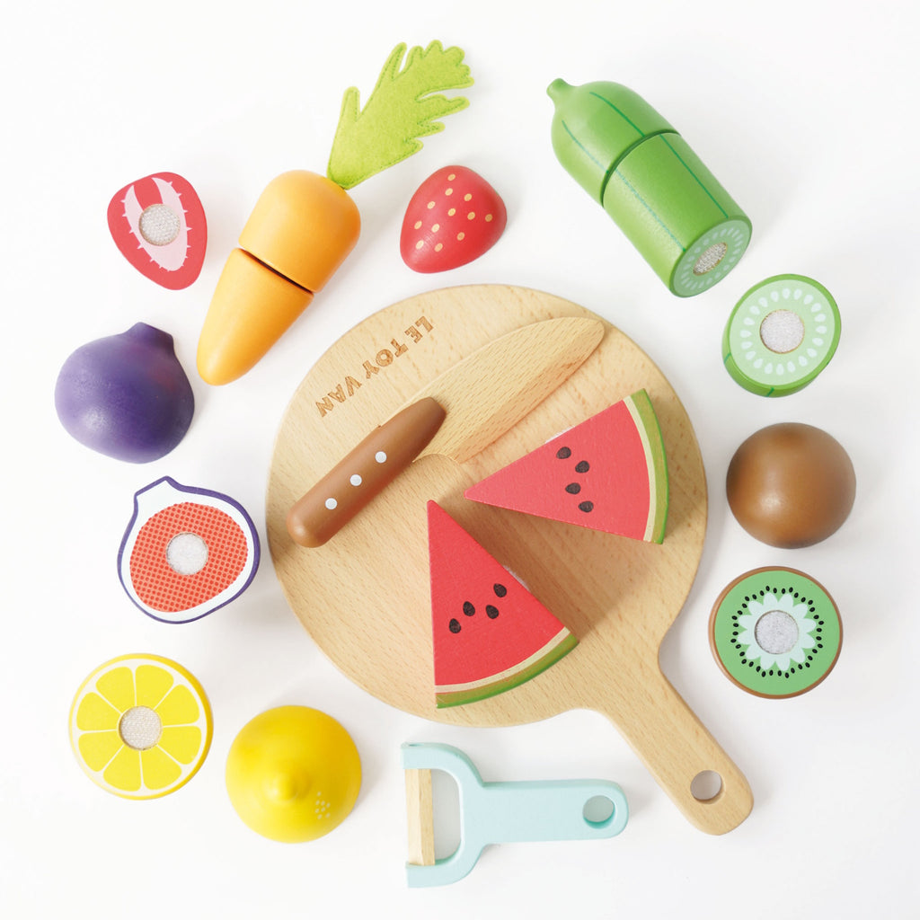 Wooden toy fruits and vegetables, including watermelon, kiwi, and carrot on a cutting board, with a toy knife and peeler, evoke playful cooking.