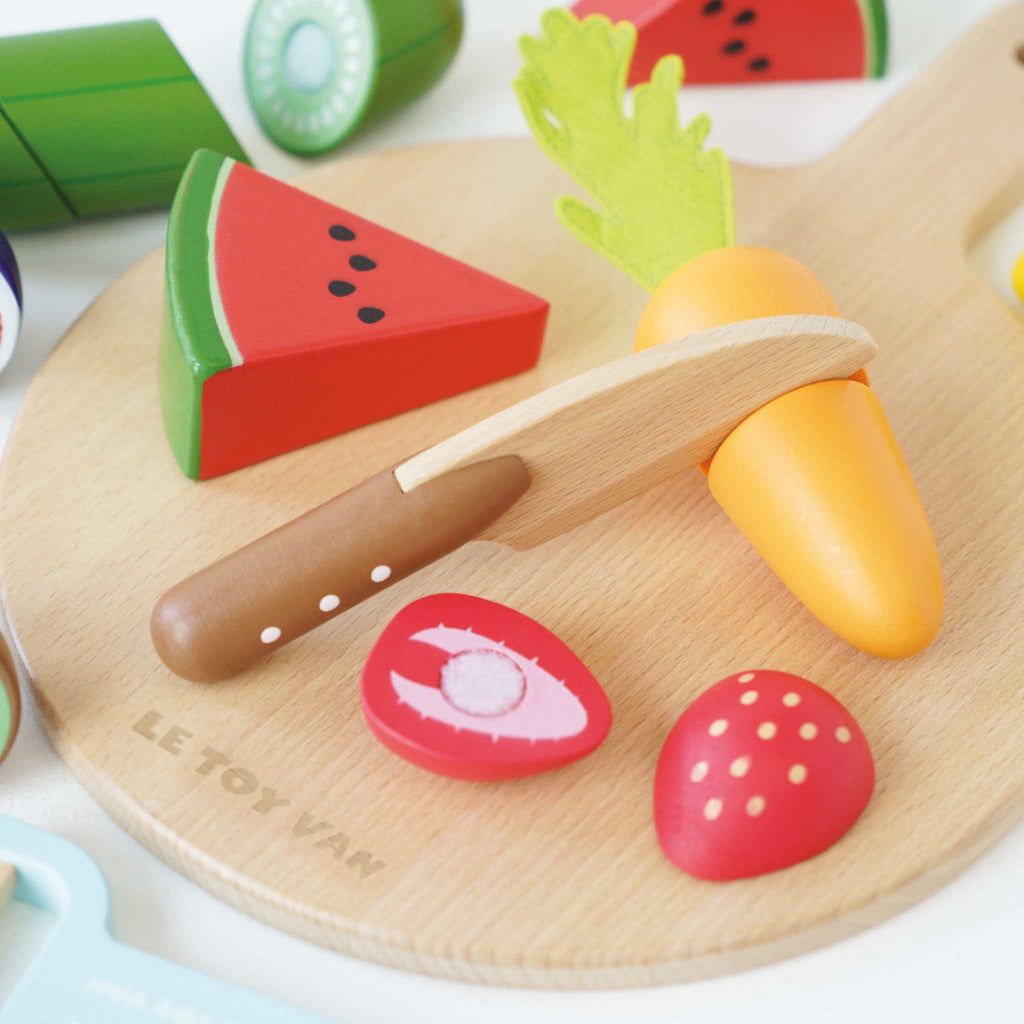 Wooden toy fruits and vegetables, including watermelon, kiwi, and carrot on a cutting board, with a toy knife and peeler, evoke playful cooking.
