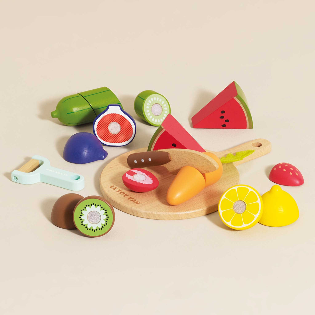 Wooden toy fruits and vegetables, including watermelon, kiwi, and carrot on a cutting board, with a toy knife and peeler, evoke playful cooking.