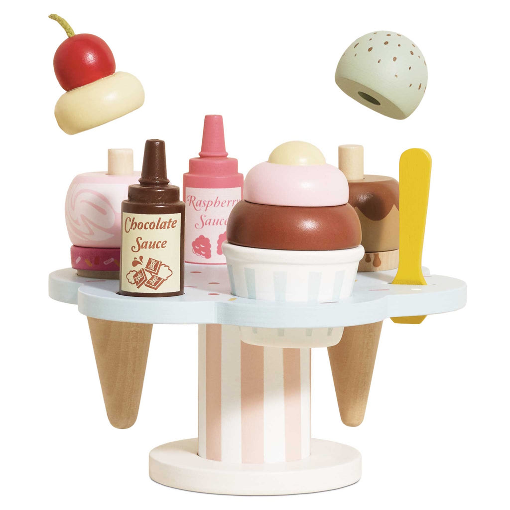 Wooden toy ice cream set with scoops, cones, and sauce bottles on a pastel stand. A cherry and mint scoop hover above, evoking playful imagination.