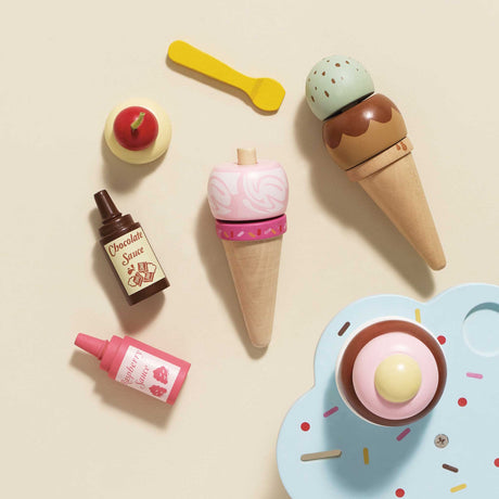 Top-down view of wooden play ice cream cones, scoops, toppings, and sauce bottles on a neutral background.