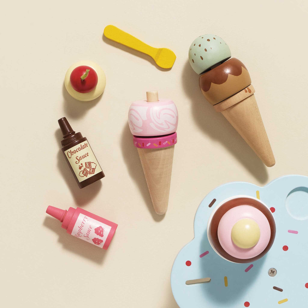 Wooden toy ice cream set with cones, scoops, a pink spoon, chocolate and raspberry sauce bottles, and a toy cupcake on a sprinkle tray. Playful and colorful.