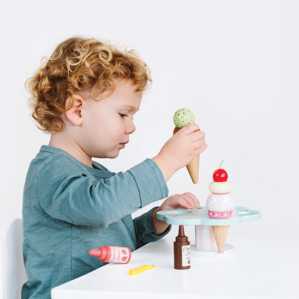 Wooden toy ice cream set with scoops, cones, and sauce bottles on a pastel stand. A cherry and mint scoop hover above, evoking playful imagination.