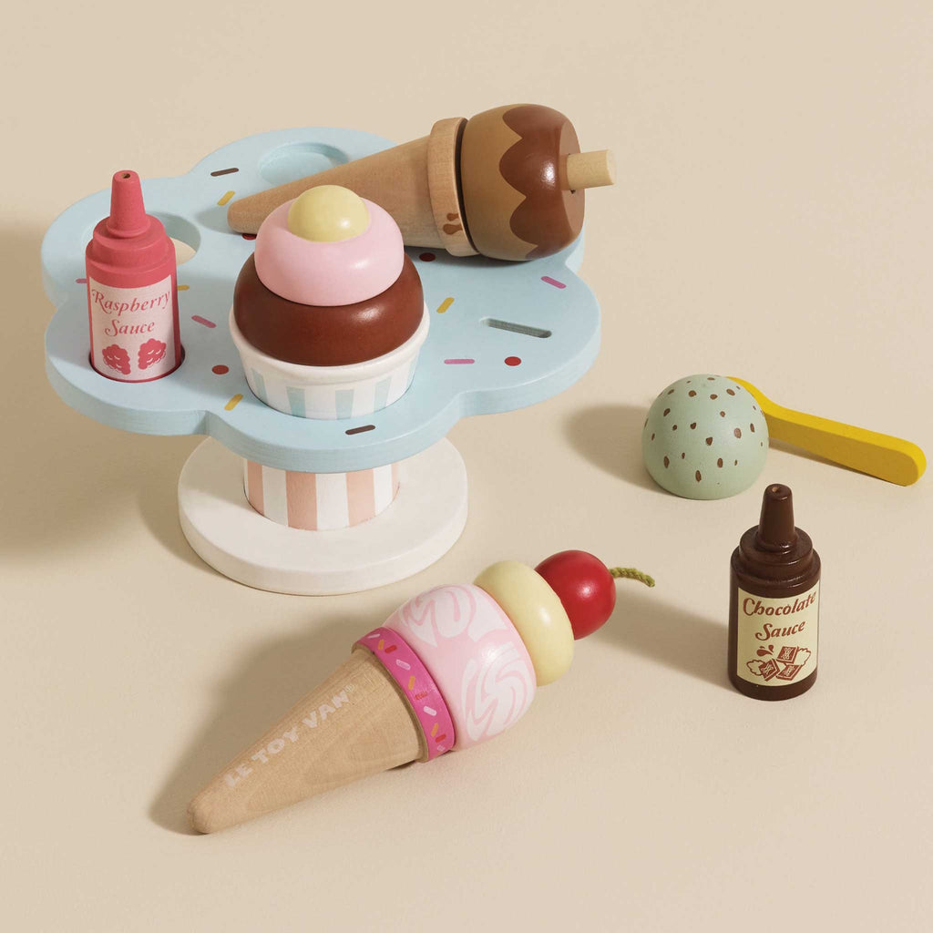 Wooden toy ice cream set with scoops, cones, and sauce bottles on a pastel stand. A cherry and mint scoop hover above, evoking playful imagination.