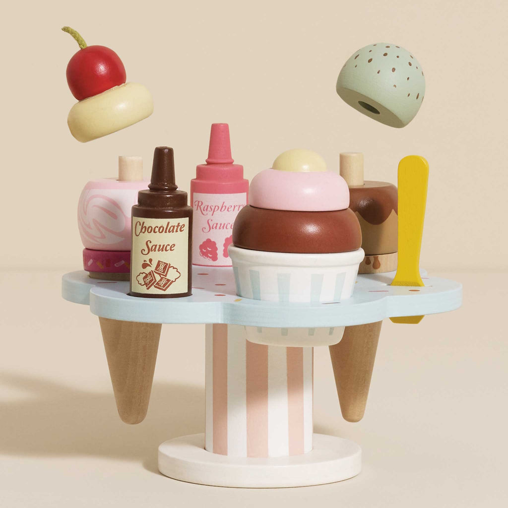Wooden toy ice cream set with scoops, cones, and sauce bottles on a pastel stand. A cherry and mint scoop hover above, evoking playful imagination.