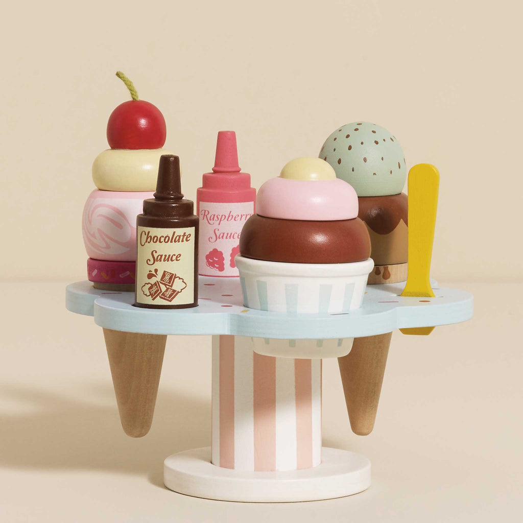 Wooden toy ice cream set with scoops, cones, and sauce bottles on a pastel stand. A cherry and mint scoop hover above, evoking playful imagination.