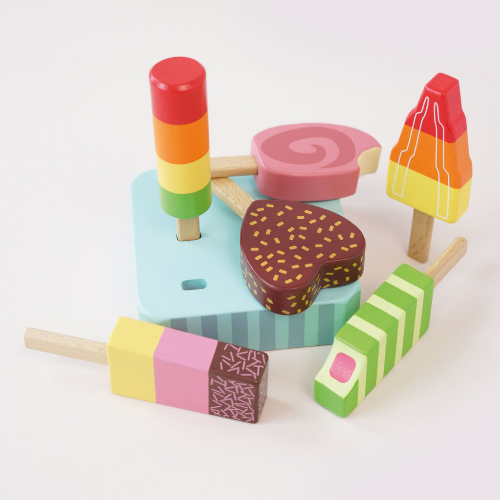 Six colorful wooden toy ice creams on sticks are arranged on a striped stand. They vary in shapes and vibrant designs, exuding a playful and whimsical tone.